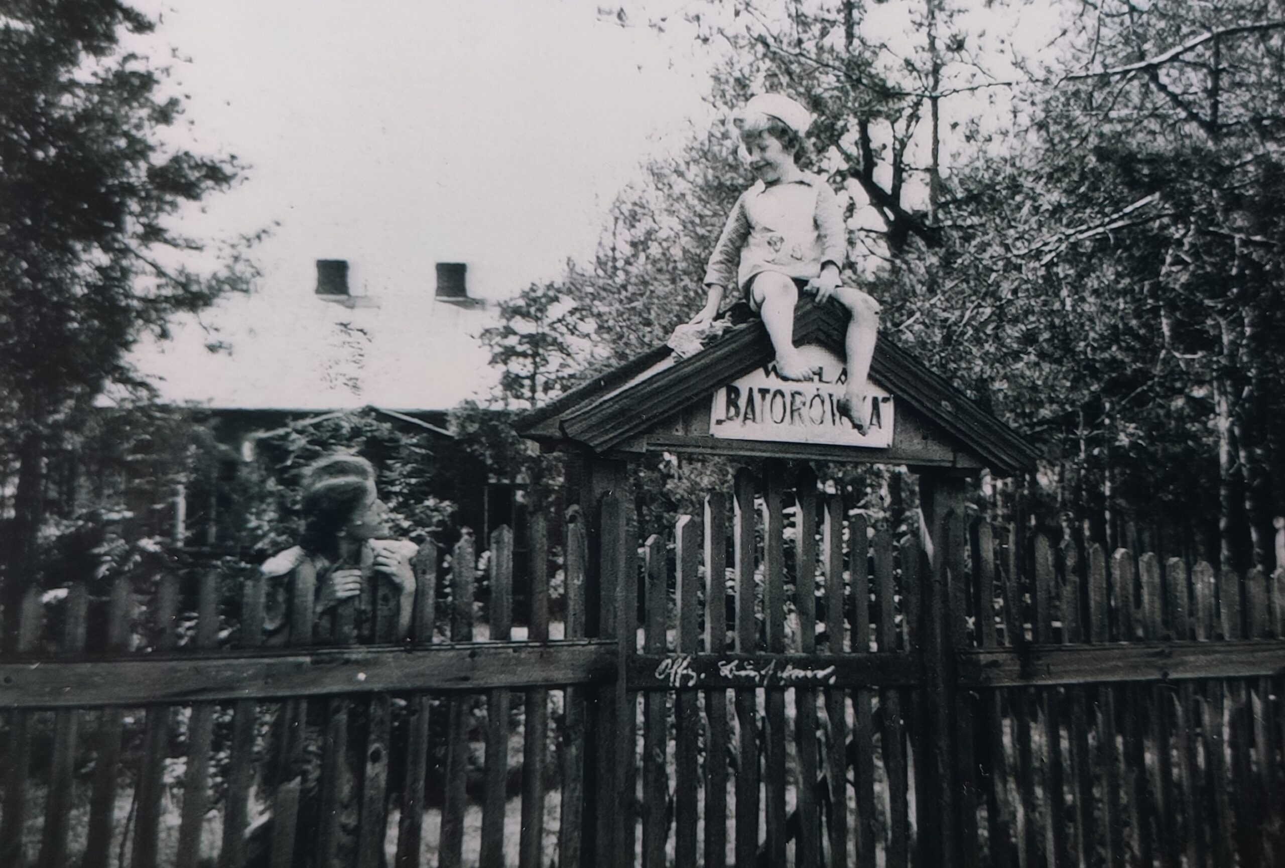 Drewniane ogrodzenie przed Willą Batorówka, nad którym widnieje napis Batorowka oraz siedzi mały chłopiec spoglądający na kobietę po lewej stronie.