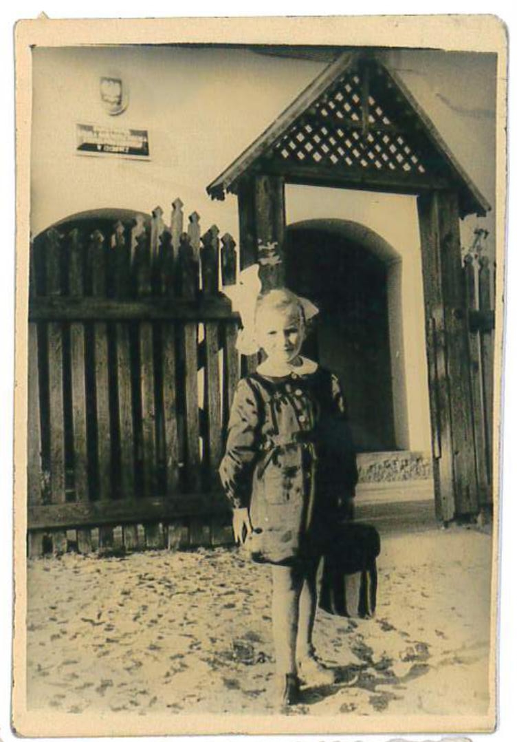 Barbara Ciszewska-Stankiewicz w czasach szkoły podstawowej. Czarno-białe, archiwalne zdjęcie przedstawia małą dziewczynkę stojącą przed drewnianym ogrodzeniem i ozdobną bramą. Dziecko ma na sobie ciemną sukienkę z jasnym kołnierzykiem oraz dużą, charakterystyczną kokardę we włosach. W dłoni trzyma małą torebkę lub teczkę, a na nogach ma jasne buciki z ciemnymi noskami. Tło stanowi budynek z łukowatym wejściem i zawieszoną na ścianie tabliczką. Całość ma sentymentalny charakter.