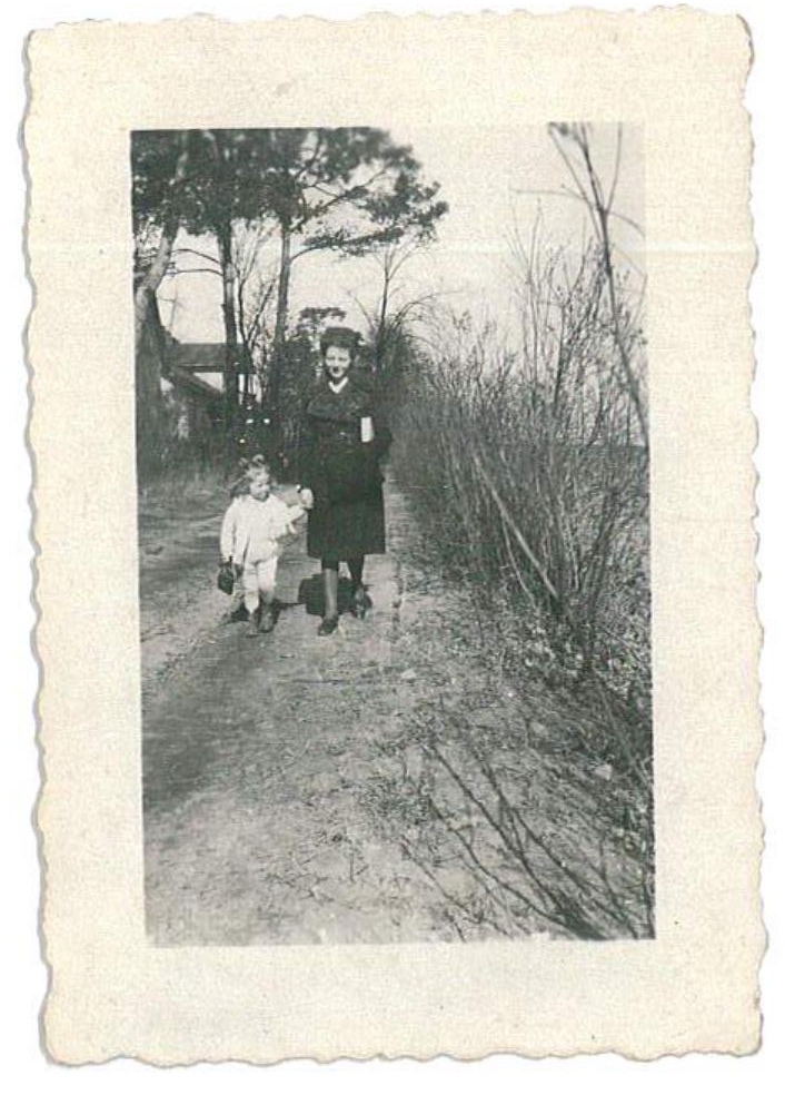 Na fotografii widać czarno-białe, nieco wyblakłe zdjęcie o postarzanym wyglądzie, z ozdobną, falowaną ramką. W centrum kadru stoi kobieta ubrana w ciemną sukienkę, trzymająca za rękę małe dziecko. Dziecko ma jasne ubranie i niewielką torebkę lub koszyczek w dłoni. Oboje idą wzdłuż wąskiej, piaszczystej ścieżki. Po obu stronach drogi rosną drzewa i krzewy — z jednej strony widać wysokie, smukłe drzewa, a z drugiej gęste, bezlistne zarośla. W tle dostrzegalny jest fragment budynku, częściowo ukryty za drzewami. Atmosfera zdjęcia jest spokojna i nostalgiczna, przywodząca na myśl dawną, rodzinną chwilę uchwyconą podczas spaceru.